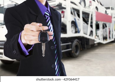 Businessman hand giving a car key with a trailer truck carrying new cars on the background
