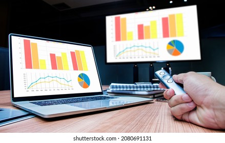 Businessman Hand Hold Remote Control Slide With Mock Up Chart Slide Show Presentation On Display Laptop And Television In Meeting Room