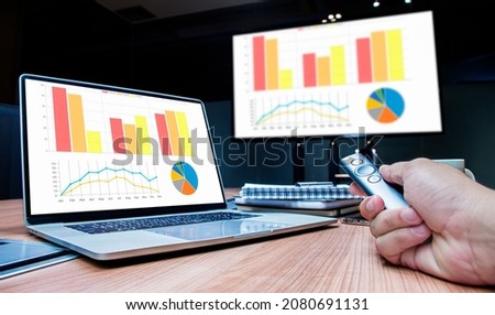 Businessman hand hold remote control slide with mock up chart slide show presentation on display laptop and television in meeting room