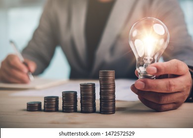 Businessman Hand Holding Lightbulb With Coins Stacking On Desk.saving Energy And Money Concept.