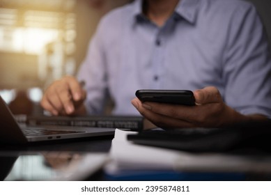 Businessman hand holding mobile smartphone on a table with a laptop at office..	 - Shutterstock ID 2395874811
