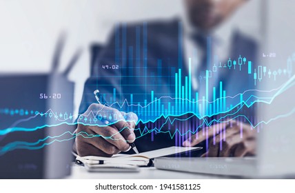 Businessman hand with pen writing in notebook, laptop on the table and double exposure of stock market changes, office interior. Blue and violet graph, growing numbers and candlesticks - Shutterstock ID 1941581125