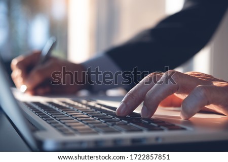 Businessman hand working on laptop computer, typing keyboard, writing on notebook planning his work in modern office, close up. Project manager, corporate man proofing business report on desk.