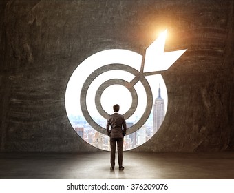 A businessman with hands in pockeys standing in front of a target through which he can see New York. Back view. Black background. Concept of achieving a goal. - Shutterstock ID 376209076