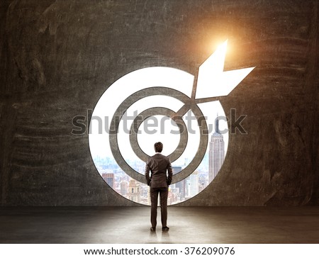 A businessman with hands in pockeys standing in front of a target through which he can see New York. Back view. Black background. Concept of achieving a goal.