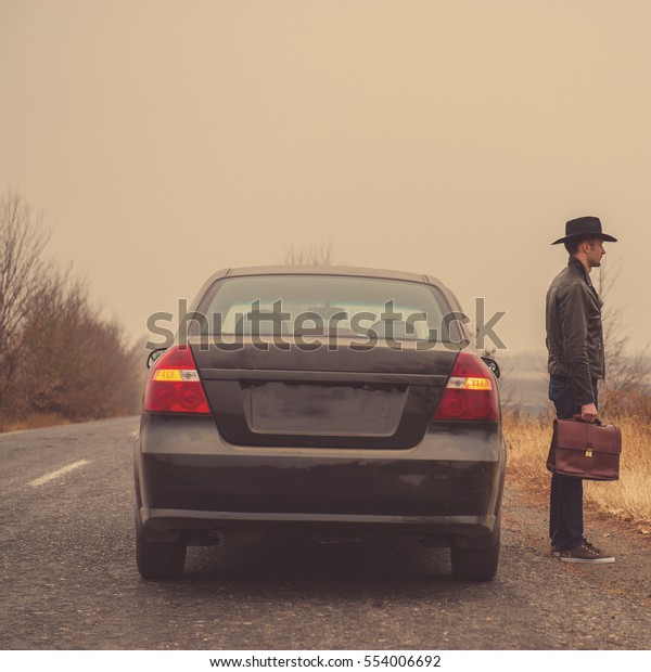 Businessman in a hat with a briefcase in his hand\
standing near a car