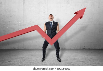 Businessman holding a big red line graph with an upturned arrow and trying to raise it up with his muscular arms. Working hard to increase incomes. Making efforts to grow profits. Improving business
