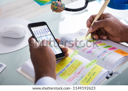 Businessman Holding Cellphone With Gantt App And Calendar Writing Schedule In Diary With Pen