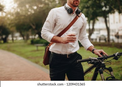 Businessman Holding A Coffee Cup And Walking To Office Taking His Bicycle Along. Man Enjoying Coffee While Walking To Work Along With His Bike.