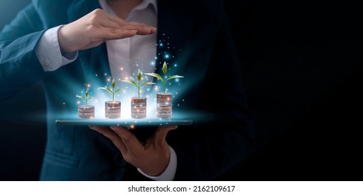 A businessman holding a coin on tablet with a tree that grows and a tree that grows on a pile of money. The idea of maximizing the profit from the business investment. - Shutterstock ID 2162109617