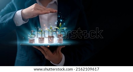 A businessman holding a coin on tablet with a tree that grows and a tree that grows on a pile of money. The idea of maximizing the profit from the business investment.