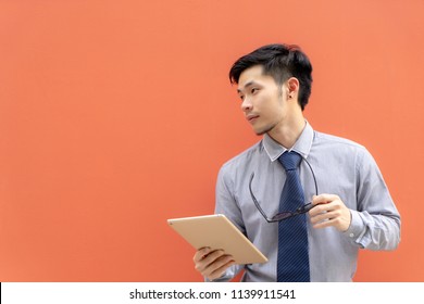 Businessman holding digital tablet computer on background with copy space.Happy young Asian businessman in shirt and tie working on digital tablet against orange background.