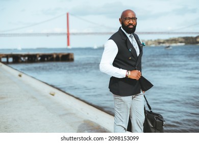 A Businessman Holding A Messenger Bag Near A River, A Red Suspension Bridge In A Blurred Background. A Bald Bearded Black Guy In A Suit Standing On The Pier Looking Away, A Copy Space Area On The Left