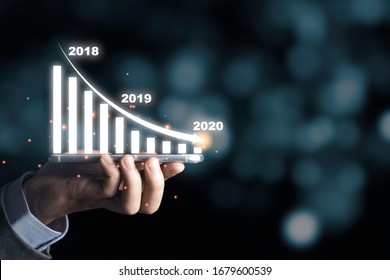 Businessman Holding Mobile Phone With Infographic Graph And Arrow Down Trend In 2020 Economic Crisis.It Is Symbol Of Business Investment Down From Trade War And Coronavirus (COVID 19).