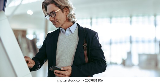 Businessman Holding Mobile Phone Using Self Service Check In Machine At International Airport. Male Passenger Doing Self Check In.