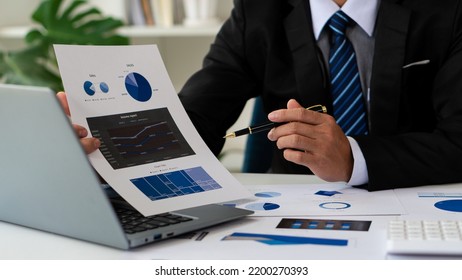 Businessman Holding Pen And Pie Chart Document Showing Company Financial Data He Sits In A Private Office With A Laptop Analyzing Financial Data In Chart Form. Financial Concept