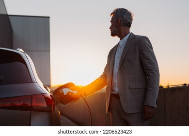 Businessman, holding power supply and charging his electric car during sunset. Concept of ecology tranport.