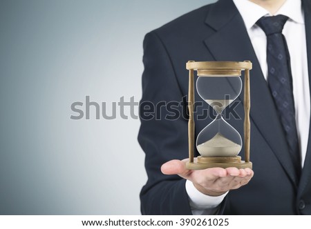 Businessman holding sand glass on palm. Sand running. Grey background. Concept of time.