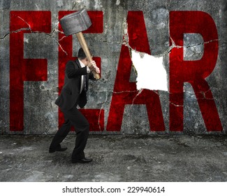 businessman holding sledgehammer hitting red FEAR word on concrete wall with large blank hole, overcoming fear concept.