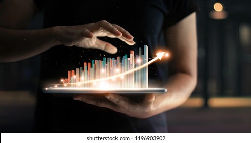 businessman holding tablet and showing a growing virtual hologram of statistics, graph and chart with arrow up on dark background. Stock market. Business growth, planing and strategy concept. 