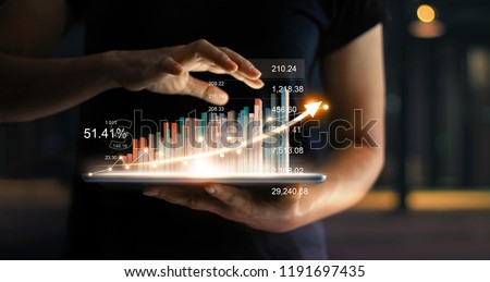 Businessman holding tablet and showing a growing virtual hologram of statistics, graph and chart with arrow up on dark background. Stock market. Business growth, planing and strategy concept. 