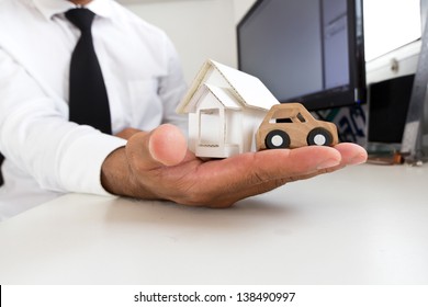 Businessman Holding A Wooden Car And Home On A Table.