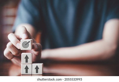 Businessman holding wooden cube with target board icon and arrow on wooden table. Goals and planning for success in marketing business, achieve the objective concept. Closeup and free copy space - Shutterstock ID 2055533993
