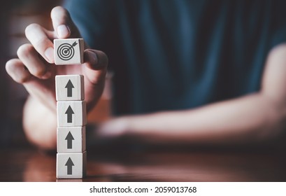 Businessman Holding Wooden Cube With Target Board Icon And Arrow On Wooden Table. Goals And Planning For Success In Marketing Business, Achieve The Objective Concept. Closeup And Free Copy Space