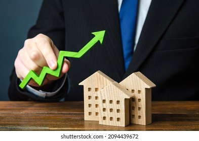 Businessman holds an arrow up over wooden houses. The concept of increasing the cost of housing. High demand for real estate. The growth of rent and mortgage rates. Sale of apartments.