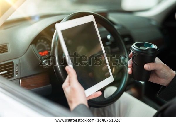 businessman holds coffee in a papper cup and reads\
news from his tablet while sitting in his car with open window,\
multitasking, big city\
life