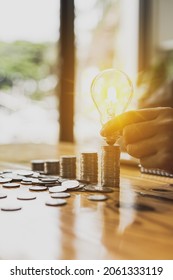 Businessman holds a glowing light bulb on top of the highest pile of coins, Placing coins in a row from low to high is comparable to saving money to grow more. Money saving ideas for investing in fund - Shutterstock ID 2061333119