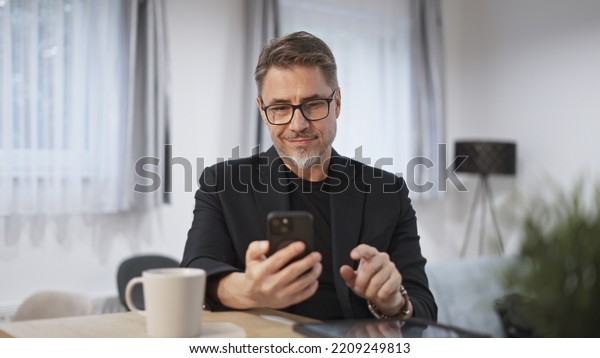 Businessman at home, using phone,\
checking newsfeed. Home office, browsing internet. Portrait of\
happy, mature age, middle age, mid adult man in 50s,\
smiling.