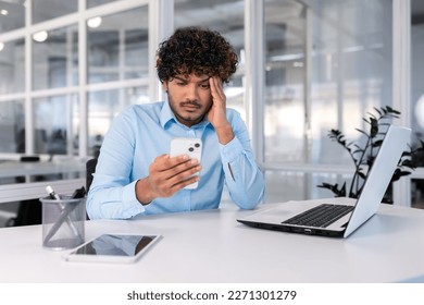 Businessman inside the office at the workplace is not satisfied with the work of the phone, the man is upset holding a smartphone in his hands, hispanic man is sitting at the table with a laptop.