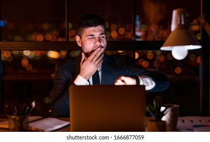 Businessman At Laptop Yawning Checking Time Looking At Watch Working Late At Night In Modern Office. Selective Focus