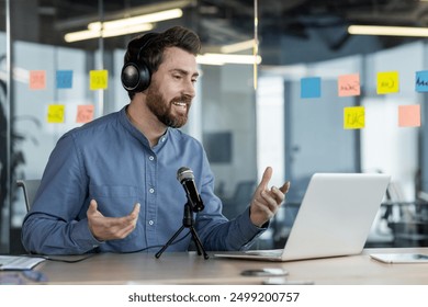 Businessman leading virtual meeting using laptop, microphone, headphones in modern office. Professional engaging in online conference with sticky notes on glass wall in background. - Powered by Shutterstock