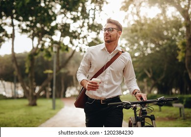 Businessman listening to music and enjoying coffee while walking to office with his bike. Man enjoying his commute to office walking with his bicycle and a coffee cup in hand. - Powered by Shutterstock