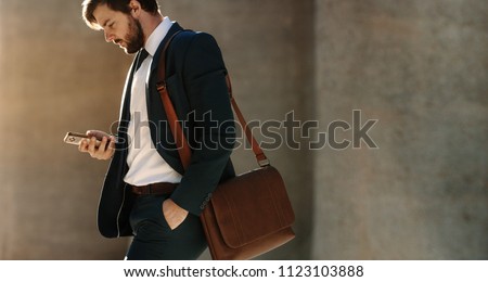 Businessman looking at his mobile phone while walking on street to office. Busy office going man commuting to office carrying his office bag and using mobile phone.