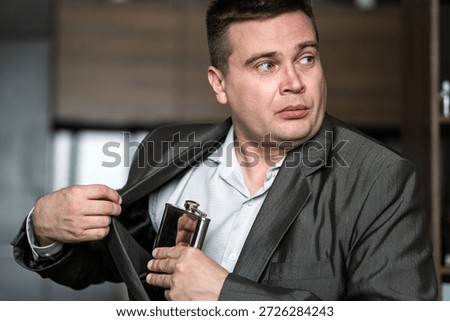 Businessman looking scared while taking a hip flask out of his jacket at work, hiding alcohol addiction issues of alcoholism in the workplace. The man hides a flask of rum in his jacket pocket.