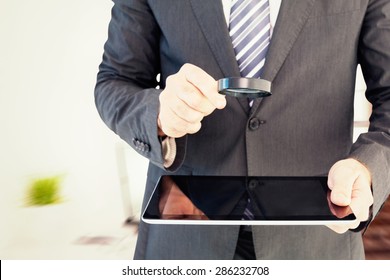 Businessman looking at tablet with magnifying glass against laptop on desk with glasses and notepad - Powered by Shutterstock