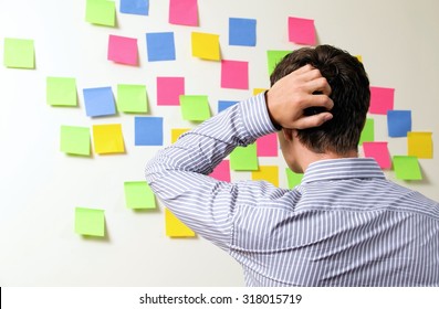 Businessman Looking At Wall Of Sticky Notes With Hands Behind Head