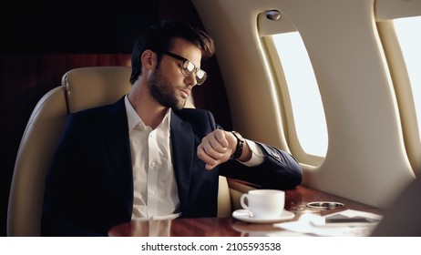 Businessman looking at wristwatch near coffee and cellphone in private plane