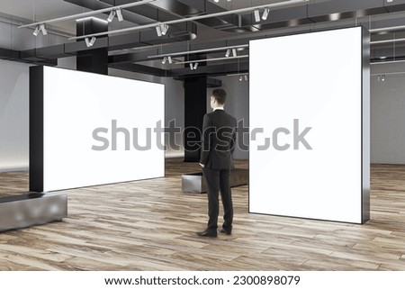Businessman looks at blank white partitions with place for advertising poster or marketing campaign in stylish gallery hall with metallic benches on wooden floor and grey wall background, mockup