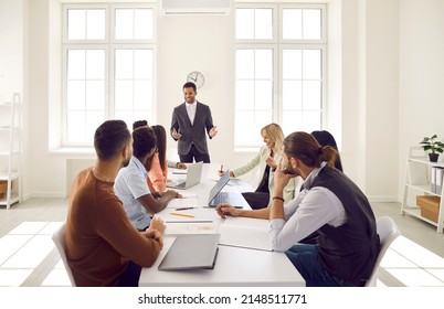 Businessman Makes Suggestions To Colleagues, Conducts Business Training Or Discusses Financial Results At Corporate Briefing. Diverse Employees Sitting At Long Table In Office At Business Meeting.