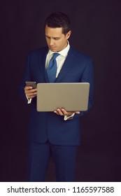 Businessman. Mobile Phone While Working On Laptop On Black Background. Man Or Manager Wearing Formal Blue Suit. Technology For Business. Messaging. Fashion And Dress Code