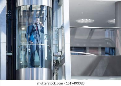 Businessman In Modern Glass Elevator Talking By The Phone And Holding Briefcase