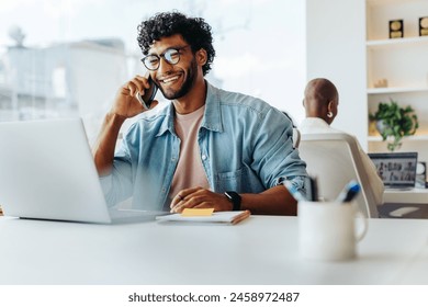 Businessman in a modern office, curly-haired, wearing glasses, works on a laptop, smiling while talking on the phone. Surrounded by technology and a smartphone, he exudes professionalism and success.