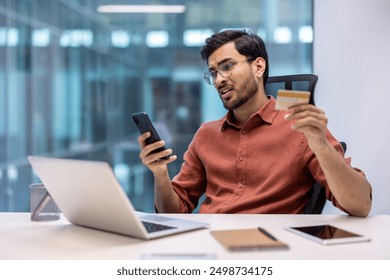 Businessman in modern office holding credit card, looking concerned about phone fraud. Laptop, notebook, and tablet on desk. Concept of online fraud, cyber security, and technology issues