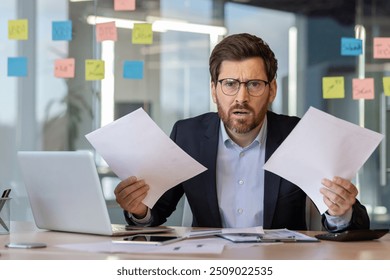 Businessman at office desk overwhelmed by work holding papers. Frustrated expression suggests stress and urgency. Laptop and phone on desk indicate multitasking in modern workplace setting. - Powered by Shutterstock