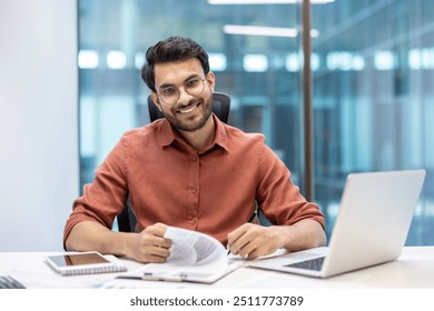 Businessman in an office environment smiling confidently while looking at documents. Laptop and phone visible, suggesting multitasking and productivity. Modern workspace background - Powered by Shutterstock