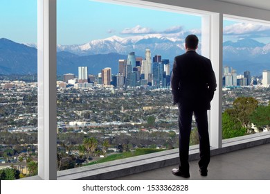 Businessman in an office looking at the view of downtown Los Angeles. The man looks like a boss or a regional manager working in California. The background is snowy San Gabriel Mountains in winter.